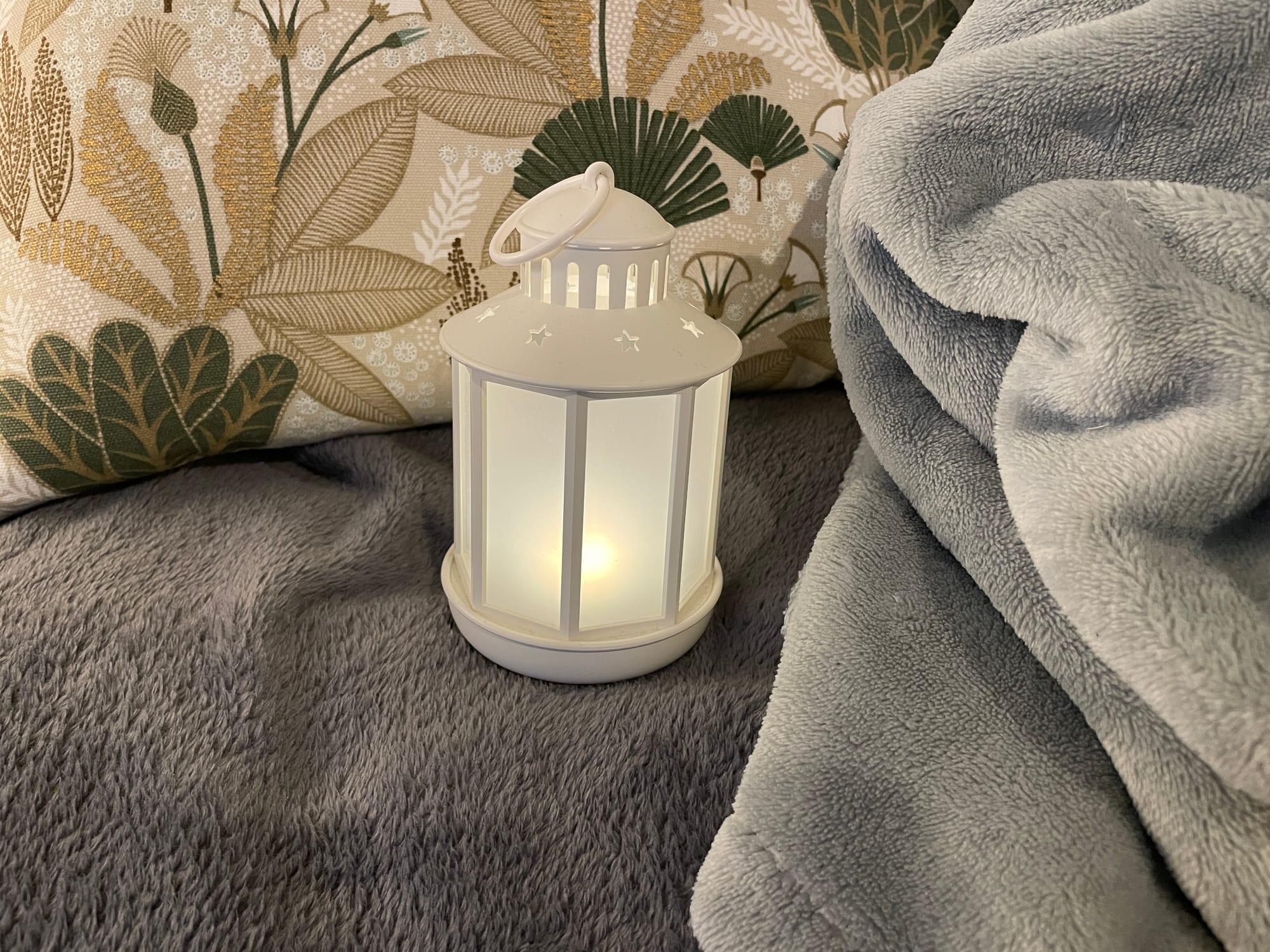 Une petite lanterne LED blanche allumée, posée à côté d'un coussin et de couvertures douces