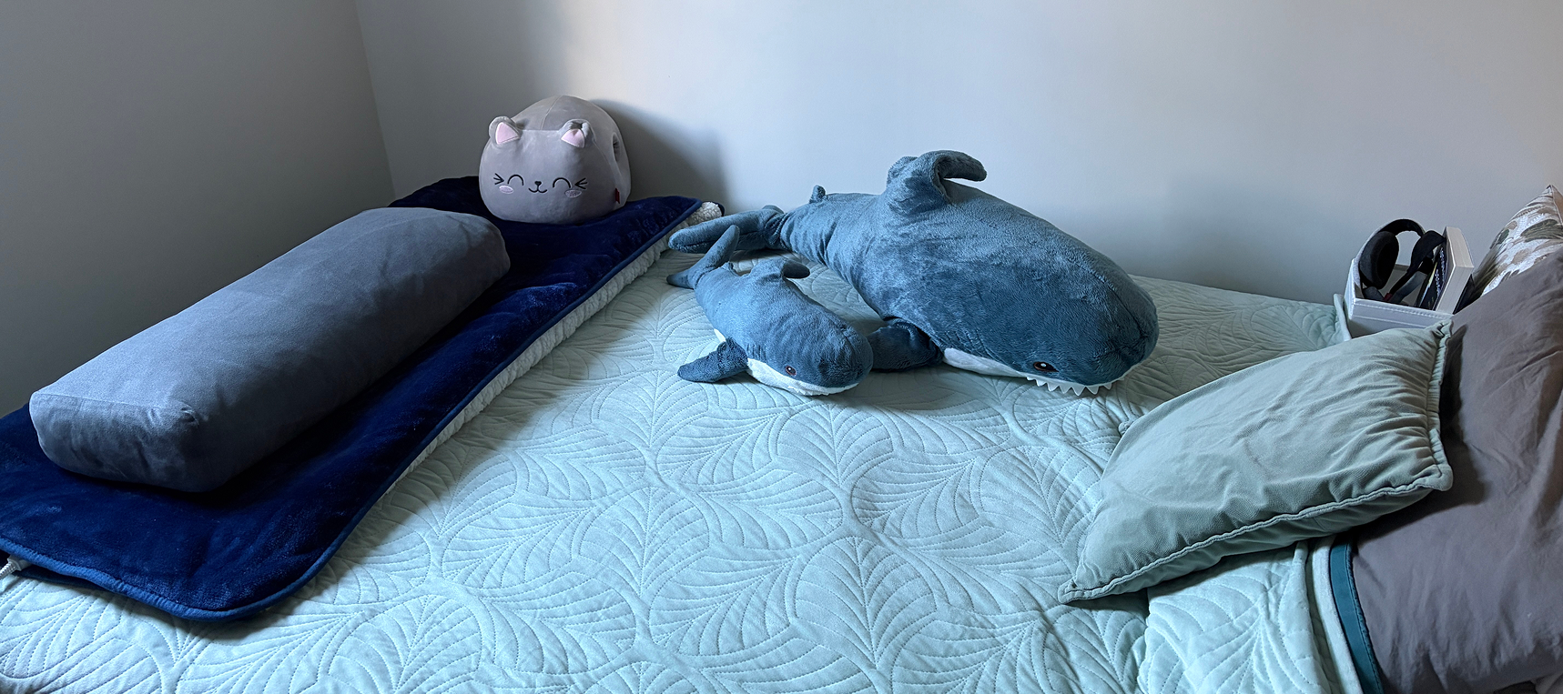 A picture of my bed with two shark plushies, a cat plushie, a yoga bolster and a some cushions, on a teal blanket.